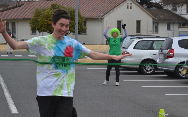 A young male runner wearing bib number 218 and a tie-dye shirt is breaking through a green finish line tape at what appears to be a sprint race event. He is smiling and has his arms outstretched as he crosses the finish line. In the background, a person wearing a bright green wig and green shirt is cheering with raised fists.