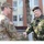 Two military personnel from different nations are shaking hands in what appears to be a formal greeting or meeting. The soldier on the left wears a US Army uniform with an Airborne tab and 25th Infantry Division patch, while the soldier on the right wears a Romanian Army uniform with a maroon beret and NATO-affiliated insignia. The setting appears to be an outdoor area near a military or administrative building.