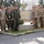 A large group of military personnel from multiple nations pose for a group photograph in front of a stone sign reading 'NATO Force Integration Unit Romania'. The personnel are wearing various camouflage uniforms representing different NATO member nations. The setting appears to be a military installation in Romania.