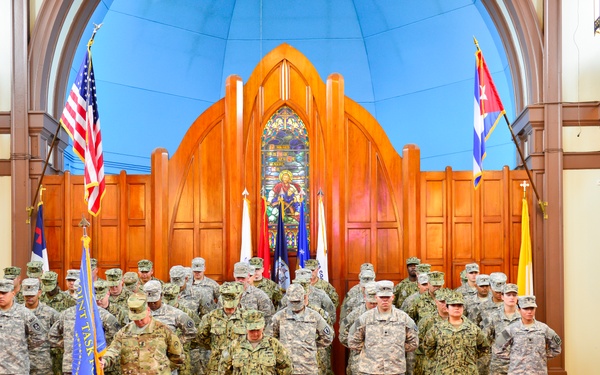 A formal military ceremony is taking place inside what appears to be a chapel or ceremonial hall, featuring a large group of uniformed service members from multiple branches standing in formation. The setting includes a prominent stained glass window, wooden Gothic-style altar backdrop, a red carpet, and multiple flags including the U.S. flag and what appears to be a Cuban flag. Two central figures appear to be conducting a guidon or unit colors ceremony on a raised platform.
