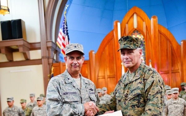 Two senior military officers are shaking hands during a formal awards ceremony, jointly holding a certificate. The officer on the left wears U.S. Air Force ABU (Airman Battle Uniform) with the name tape 'MONTEAGUDD' and a single star indicating Brigadier General rank. The officer on the right wears U.S. Marine Corps MARPAT digital camouflage with four stars visible, indicating General rank. Rows of soldiers stand at attention in the background.