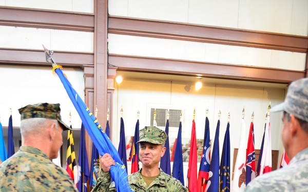 A military ceremony, likely a change of command or activation ceremony, is taking place indoors. Three service members from different branches — U.S. Marine Corps, U.S. Navy, and U.S. Air Force — are participating in a flag transfer ritual. Multiple state and organizational flags are displayed on staffs in the background.