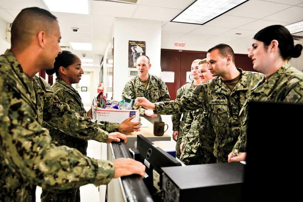 A group of U.S. Navy personnel in NWU (Navy Working Uniform) Type III camouflage are gathered around a counter or desk in what appears to be an administrative or postal office setting. One sailor is holding a USPS Priority Mail box filled with various items, suggesting a mail or care package distribution activity. The group appears engaged in a transaction or exchange, with visible name tapes and rank insignia on their uniforms.