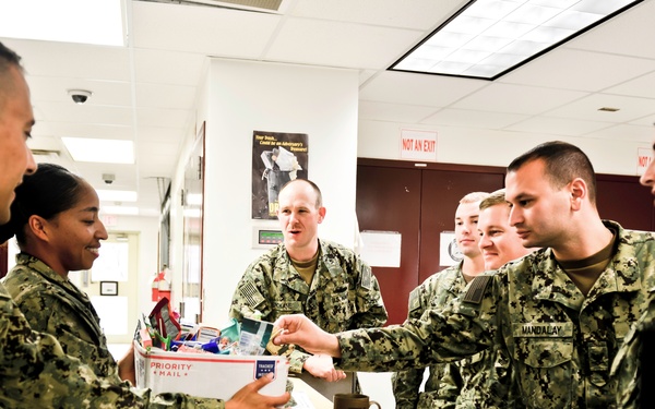 A group of U.S. Navy personnel in NWU (Navy Working Uniform) Type III camouflage are gathered around a counter or desk in what appears to be an administrative or postal office setting. One sailor is holding a USPS Priority Mail box filled with various items, suggesting a mail or care package distribution activity. The group appears engaged in a transaction or exchange, with visible name tapes and rank insignia on their uniforms.