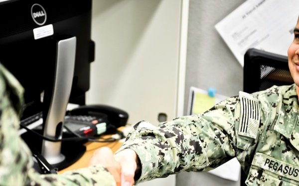 Two U.S. Navy personnel in NWU (Navy Working Uniform) digital camouflage are shaking hands in what appears to be an office or administrative setting. The servicemember on the right, whose name tape reads 'PEGASUS,' is seated at a desk and smiling. A U.S. flag patch is visible on the right shoulder of the seated individual, indicating active U.S. military service.