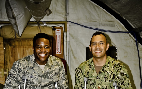 Two U.S. military service members, one from the U.S. Air Force and one from the U.S. Navy, are posing together while using crutches inside what appears to be a military field tent or temporary structure. Both men are smiling broadly at the camera despite their apparent injuries. The Navy service member's right foot/ankle appears to be bandaged or in a medical boot.