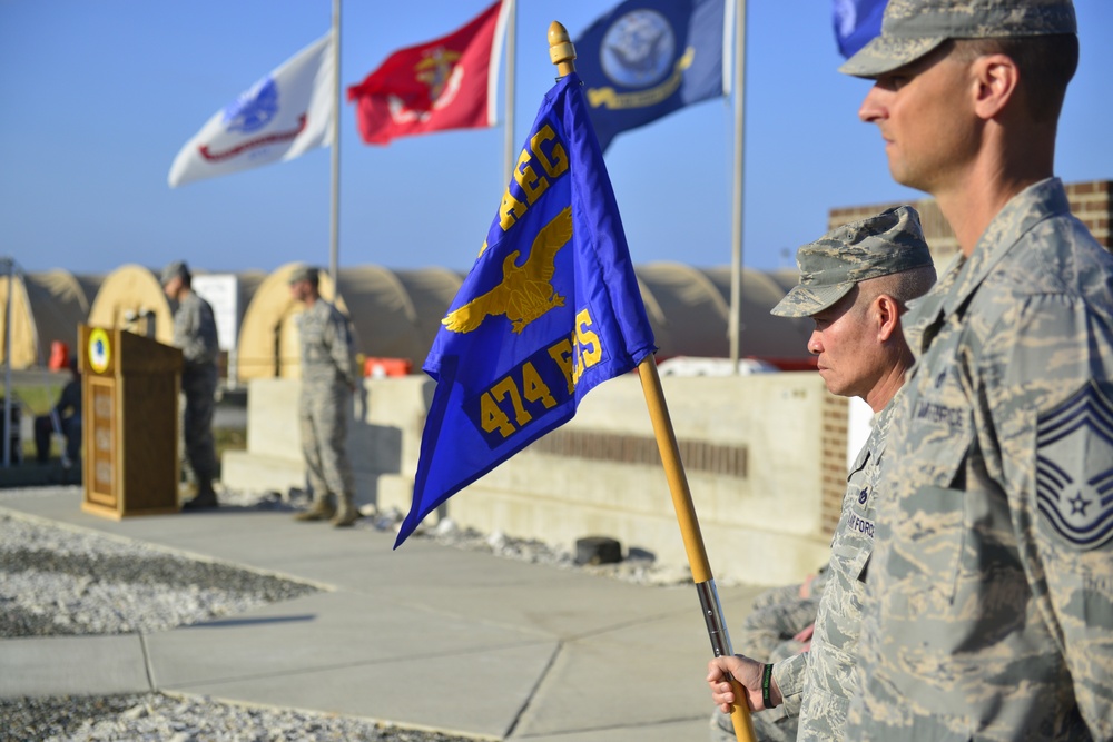 A military ceremony is taking place outdoors, likely at an air base. Personnel in US Air Force ABU/ACU camouflage uniforms are standing at attention, with one holding a blue guidon flag marked '474 ES' and featuring a gold eagle emblem. In the background, a speaker addresses attendees from a podium, with multiple military branch flags flying overhead.