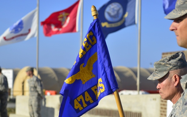A military ceremony is taking place outdoors, likely at an air base. Personnel in US Air Force ABU/ACU camouflage uniforms are standing at attention, with one holding a blue guidon flag marked '474 ES' and featuring a gold eagle emblem. In the background, a speaker addresses attendees from a podium, with multiple military branch flags flying overhead.