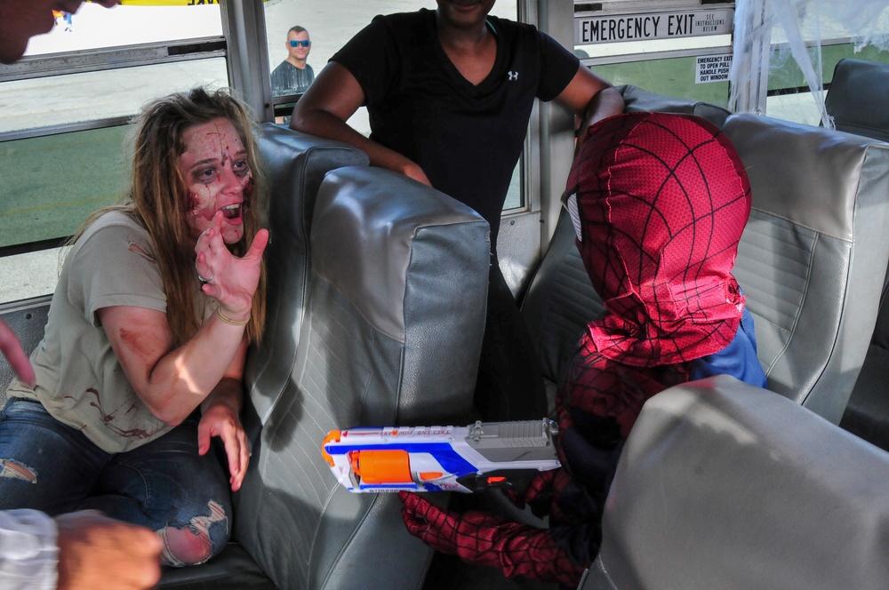 The image shows what appears to be a Halloween or costume event taking place inside a bus. A woman wearing zombie-style makeup and costume with simulated blood sits across from a child dressed in a Spider-Man costume holding a Nerf gun. The scene is clearly theatrical and playful in nature.