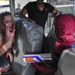 The image shows what appears to be a Halloween or costume event taking place inside a bus. A woman wearing zombie-style makeup and costume with simulated blood sits across from a child dressed in a Spider-Man costume holding a Nerf gun. The scene is clearly theatrical and playful in nature.