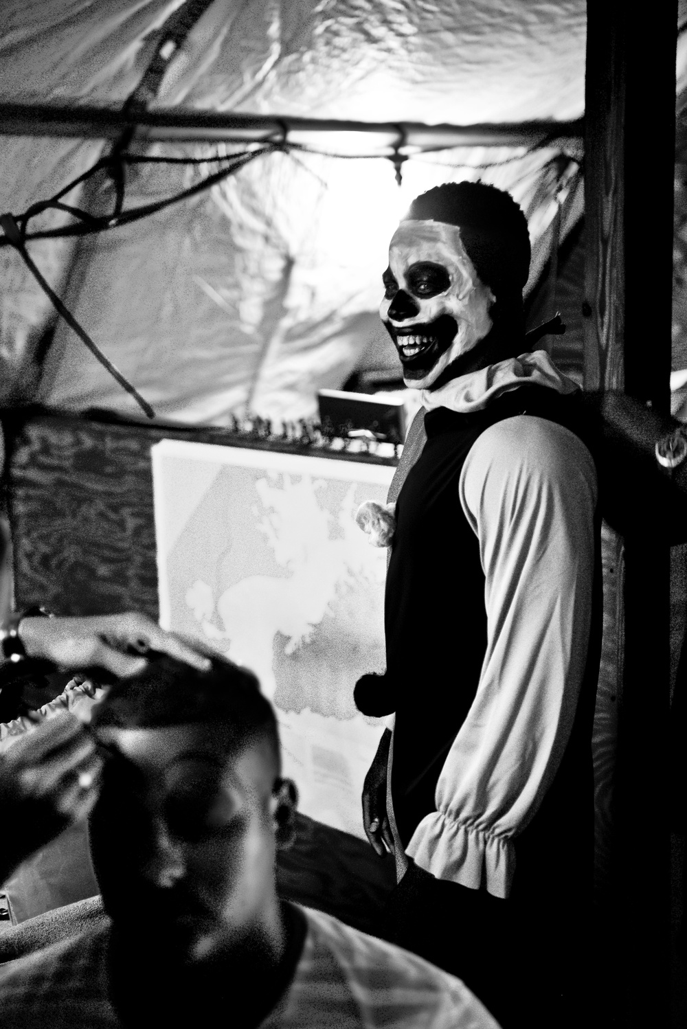 A black and white photograph showing a person wearing skull/skeleton face paint and a costume with a ruffled collar, smiling broadly. The setting appears to be a Halloween haunted house or themed event, with decorative elements including fake cobwebs and draped plastic sheeting. A second person is partially visible in the foreground.