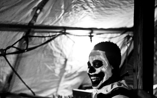 A black and white photograph showing a person wearing skull/skeleton face paint and a costume with a ruffled collar, smiling broadly. The setting appears to be a Halloween haunted house or themed event, with decorative elements including fake cobwebs and draped plastic sheeting. A second person is partially visible in the foreground.