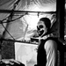 A black and white photograph showing a person wearing skull/skeleton face paint and a costume with a ruffled collar, smiling broadly. The setting appears to be a Halloween haunted house or themed event, with decorative elements including fake cobwebs and draped plastic sheeting. A second person is partially visible in the foreground.
