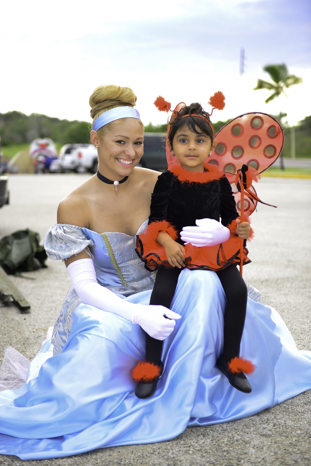 A young woman dressed as Cinderella in a light blue ball gown with white gloves and a blue headband sits outdoors holding a small child on her lap. The child is wearing a ladybug costume complete with red and black wings, antenna headband, and a decorative wand. The setting appears to be an outdoor event, likely Halloween or a themed celebration, with vehicles and trees visible in the background.
