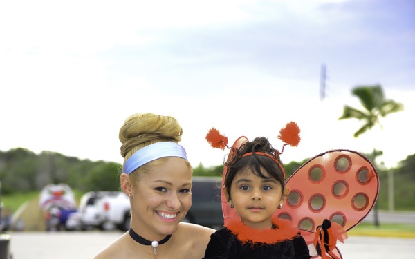 A young woman dressed as Cinderella in a light blue ball gown with white gloves and a blue headband sits outdoors holding a small child on her lap. The child is wearing a ladybug costume complete with red and black wings, antenna headband, and a decorative wand. The setting appears to be an outdoor event, likely Halloween or a themed celebration, with vehicles and trees visible in the background.