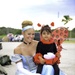 A young woman dressed as Cinderella in a light blue ball gown with white gloves and a blue headband sits outdoors holding a small child on her lap. The child is wearing a ladybug costume complete with red and black wings, antenna headband, and a decorative wand. The setting appears to be an outdoor event, likely Halloween or a themed celebration, with vehicles and trees visible in the background.