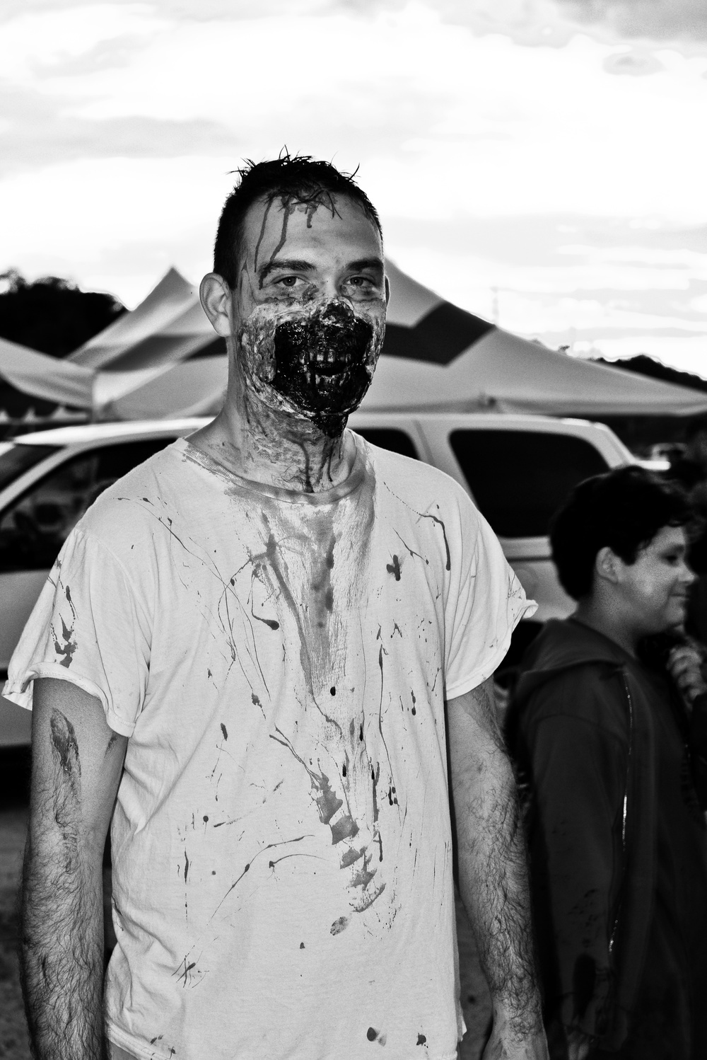 A man wearing elaborate zombie Halloween costume makeup stands facing the camera at what appears to be an outdoor event. His face features prosthetic gore makeup around the mouth area with simulated blood/dark liquid dripping down his face and onto his white t-shirt. A child in a dark costume is visible in the background near event tents and parked vehicles.