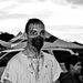 A man wearing elaborate zombie Halloween costume makeup stands facing the camera at what appears to be an outdoor event. His face features prosthetic gore makeup around the mouth area with simulated blood/dark liquid dripping down his face and onto his white t-shirt. A child in a dark costume is visible in the background near event tents and parked vehicles.
