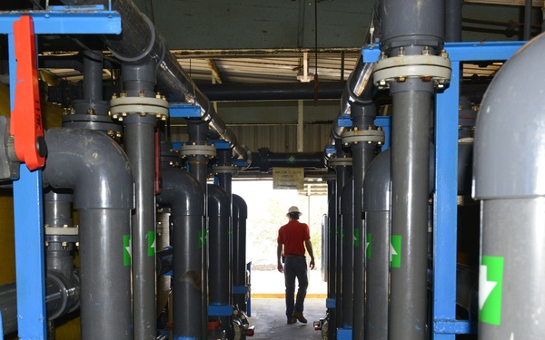 An industrial facility, likely a water treatment or filtration plant, showing rows of large cylindrical filter vessels mounted on blue steel frames with extensive gray PVC piping and valves. A worker wearing a red shirt, jeans, and a white hard hat is walking away from the camera through the narrow aisle between the equipment. The facility appears to be a semi-enclosed structure with natural light visible at the far end.