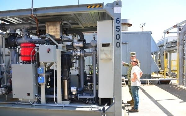 Two individuals, one in military uniform and one in civilian clothing, are inspecting an industrial equipment unit labeled '6500'. The unit appears to be a containerized mechanical or fluid processing system with visible piping, pumps, valves, and control panels. The setting is an outdoor industrial or military facility with additional structures visible in the background.