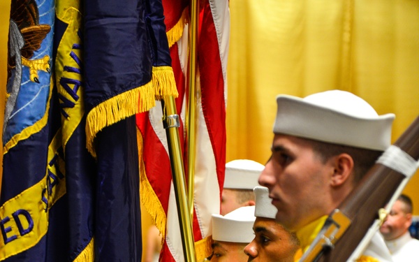 A U.S. Navy color guard is performing a ceremonial presentation in a formal indoor setting. Personnel are dressed in white dress uniforms with white gloves, carrying flags including the American flag and a U.S. Navy flag with gold fringe. The foreground sailor carries a rifle, indicating this is a formal military color guard ceremony.