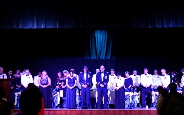 A formal military ball or gala event is taking place in a large venue. A group of approximately 20 individuals in military dress uniforms and formal evening wear are standing on an elevated stage, appearing to observe a moment of silence or prayer with heads bowed. Audience members are visible in the foreground, also standing and facing the stage.