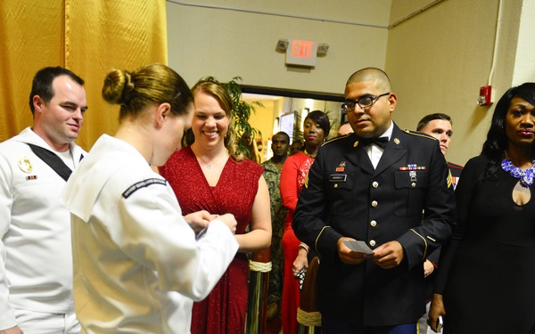 A formal military event, likely a ball or gala, where service members in dress uniforms and civilians in formal attire are being greeted or checked in at an entrance. A Navy sailor in white dress uniform appears to be checking tickets or credentials for attendees entering through a red-carpeted doorway. The scene includes personnel from multiple military branches, including Navy and Army.