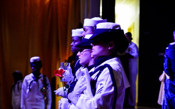 A formal military ceremony featuring U.S. Navy personnel dressed in white dress uniforms standing in a line at attention. The sailors are holding red roses and champagne glasses, suggesting a formal ball or ceremonial event. The setting appears to be a banquet hall with decorative lighting and draped curtains.