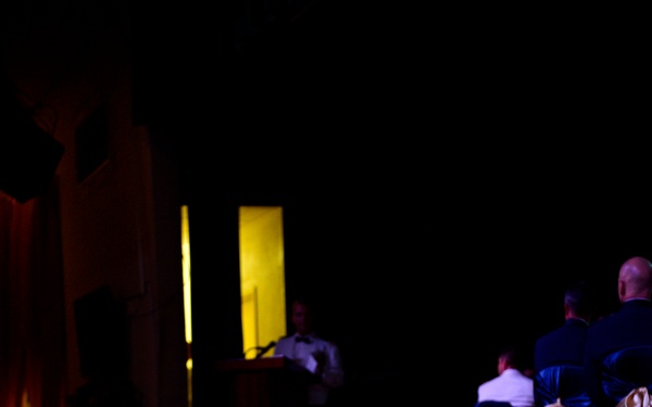 A dimly lit formal event or banquet hall features a spotlit small round table draped in a white tablecloth in the foreground. The table holds a single red rose in a glass vase, a lit candle with a yellow ribbon, a folded napkin, and a small plate. In the background, attendees in formal attire are seated near a stage where a speaker stands at a podium.