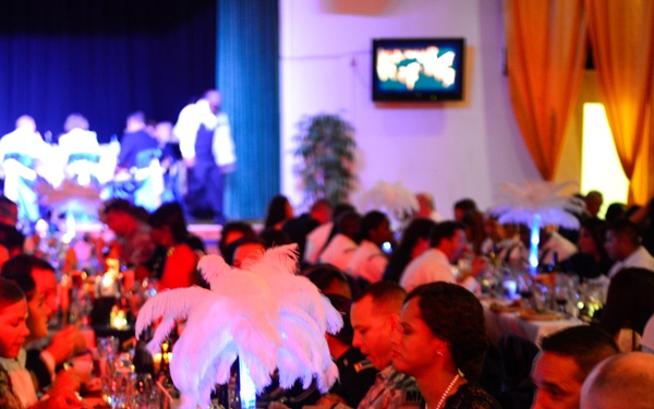A formal military banquet or ball is taking place in a large decorated venue. Attendees include uniformed military personnel with visible service ribbons and civilians in formal attire seated at long banquet tables. The event features elaborate table settings, feather centerpieces, and a live performance stage visible in the background.