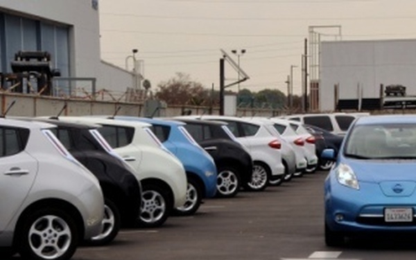 A parking lot contains a row of multiple Nissan Leaf electric vehicles in various colors including blue, white, black, and silver. One blue Nissan Leaf is positioned separately in the driving lane, facing the camera with its headlights on. The setting appears to be an industrial or commercial facility with warehouse-style buildings visible in the background.