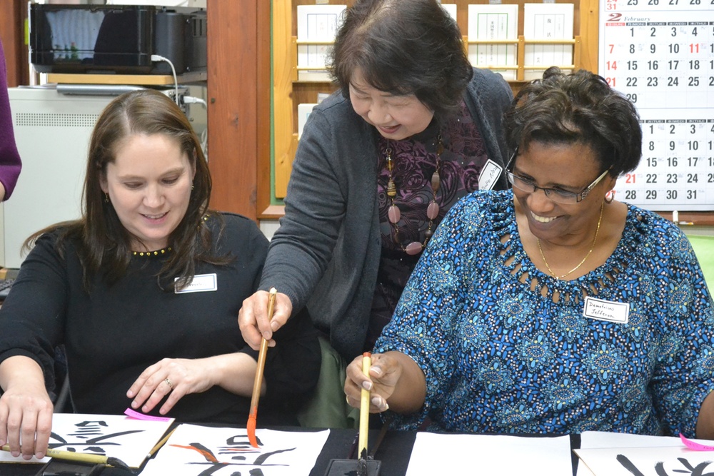 Three women are engaged in a calligraphy lesson, likely East Asian brush calligraphy. An instructor leaning over is guiding two seated students who are practicing brushwork on white paper. The setting appears to be a classroom or studio environment.