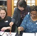 Three women are engaged in a calligraphy lesson, likely East Asian brush calligraphy. An instructor leaning over is guiding two seated students who are practicing brushwork on white paper. The setting appears to be a classroom or studio environment.