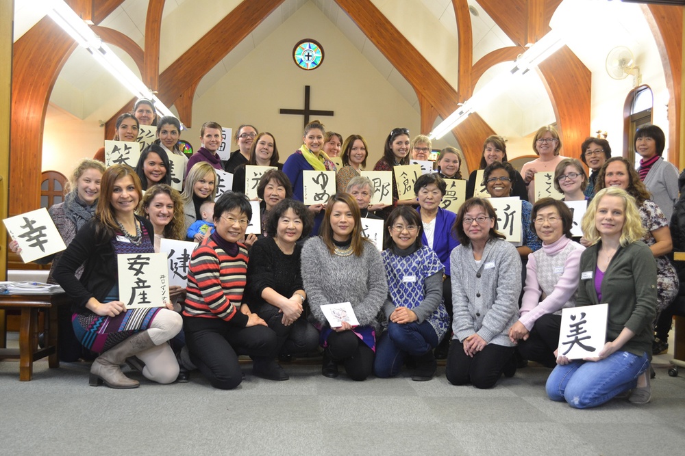 A large group of approximately 35-40 women are gathered inside a Christian church, posing for a group photo while holding handwritten Japanese calligraphy signs. The setting features wooden arched beams, a cross on the wall, and a stained glass window with a cross motif above the altar area. The group appears ethnically diverse, suggesting a multicultural women's gathering or workshop.