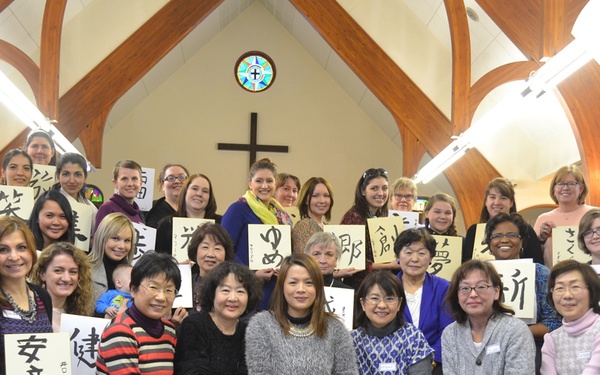A large group of approximately 35-40 women are gathered inside a Christian church, posing for a group photo while holding handwritten Japanese calligraphy signs. The setting features wooden arched beams, a cross on the wall, and a stained glass window with a cross motif above the altar area. The group appears ethnically diverse, suggesting a multicultural women's gathering or workshop.