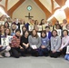 A large group of approximately 35-40 women are gathered inside a Christian church, posing for a group photo while holding handwritten Japanese calligraphy signs. The setting features wooden arched beams, a cross on the wall, and a stained glass window with a cross motif above the altar area. The group appears ethnically diverse, suggesting a multicultural women's gathering or workshop.