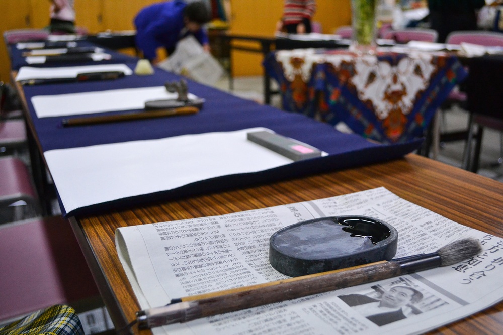 The image shows a Japanese calligraphy (shodo) class or workshop setting with multiple desks arranged in rows. Each desk is covered with a dark blue felt mat and holds white paper, calligraphy brushes, and ink stones. In the foreground, a stone ink dish (suzuri) filled with black ink and a calligraphy brush rest on a Japanese newspaper.