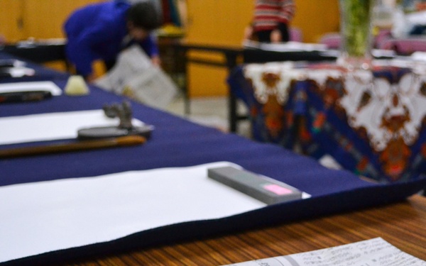 The image shows a Japanese calligraphy (shodo) class or workshop setting with multiple desks arranged in rows. Each desk is covered with a dark blue felt mat and holds white paper, calligraphy brushes, and ink stones. In the foreground, a stone ink dish (suzuri) filled with black ink and a calligraphy brush rest on a Japanese newspaper.