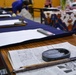 The image shows a Japanese calligraphy (shodo) class or workshop setting with multiple desks arranged in rows. Each desk is covered with a dark blue felt mat and holds white paper, calligraphy brushes, and ink stones. In the foreground, a stone ink dish (suzuri) filled with black ink and a calligraphy brush rest on a Japanese newspaper.