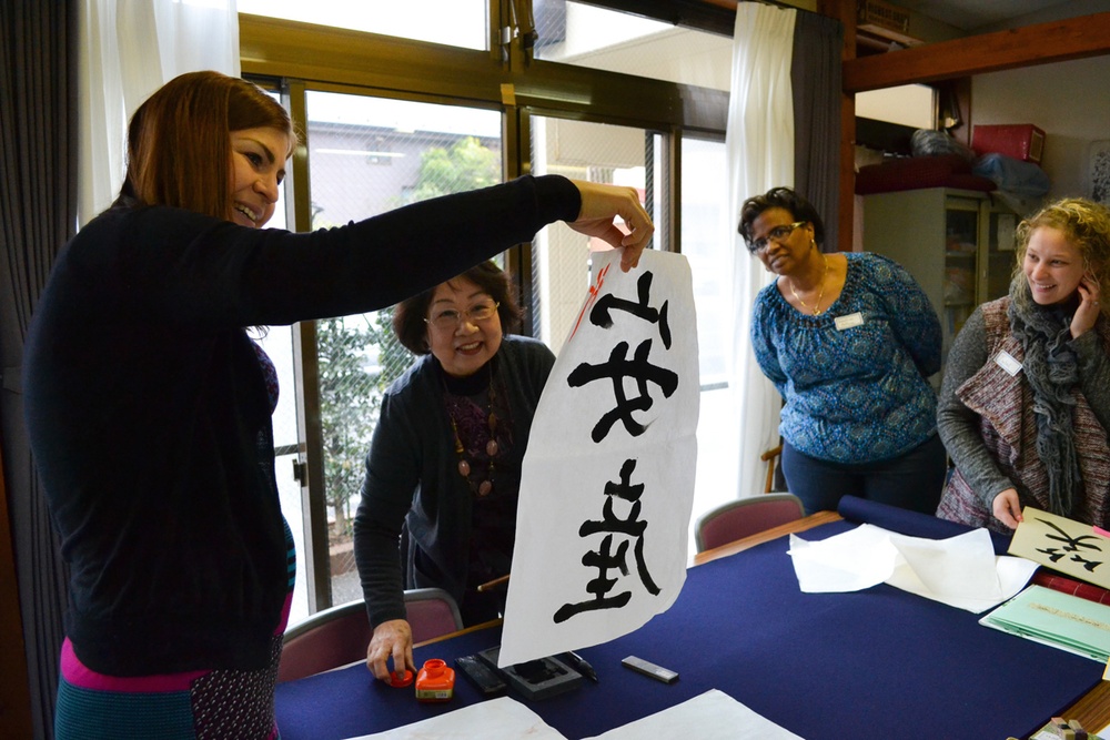 A group of four women are participating in a Japanese calligraphy workshop, likely in Japan. One woman is holding up a large piece of white paper with bold black Japanese/Chinese characters written in brush calligraphy. The setting appears to be an indoor classroom or cultural center with calligraphy supplies visible on the table.