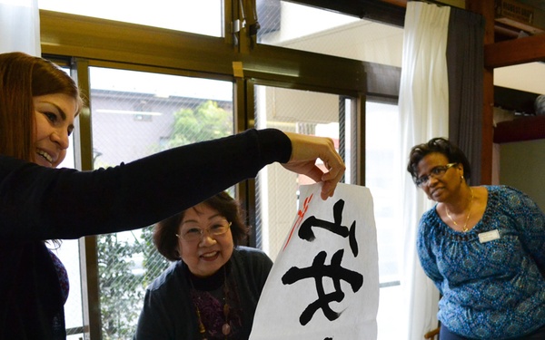 A group of four women are participating in a Japanese calligraphy workshop, likely in Japan. One woman is holding up a large piece of white paper with bold black Japanese/Chinese characters written in brush calligraphy. The setting appears to be an indoor classroom or cultural center with calligraphy supplies visible on the table.