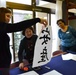 A group of four women are participating in a Japanese calligraphy workshop, likely in Japan. One woman is holding up a large piece of white paper with bold black Japanese/Chinese characters written in brush calligraphy. The setting appears to be an indoor classroom or cultural center with calligraphy supplies visible on the table.