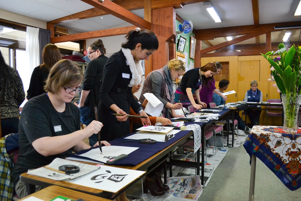A group of people, predominantly women, are participating in a Japanese calligraphy (shodo) workshop in what appears to be a traditional Japanese community room. Participants are seated or standing at long tables covered with dark blue cloth, practicing brush calligraphy on white paper. An instructor wearing a name tag and white scarf is actively guiding students at the tables.