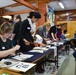 A group of people, predominantly women, are participating in a Japanese calligraphy (shodo) workshop in what appears to be a traditional Japanese community room. Participants are seated or standing at long tables covered with dark blue cloth, practicing brush calligraphy on white paper. An instructor wearing a name tag and white scarf is actively guiding students at the tables.