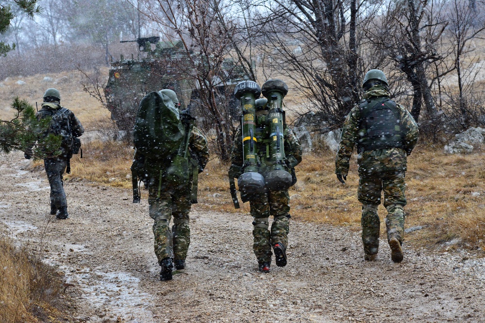 Four soldiers in camouflage uniforms are walking along a gravel path in snowy, winter conditions. The center soldier is carrying what appears to be a dual-tube man-portable missile launcher system on their back. A military armored vehicle is visible in the background among bare trees.
