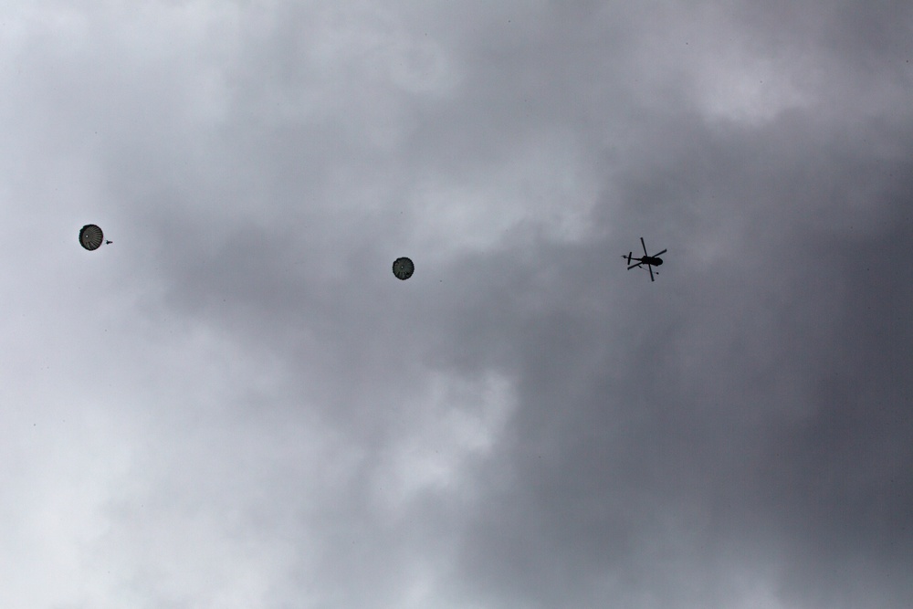 The image shows an overcast sky with two parachutes descending and a helicopter flying nearby. The silhouettes of the parachutes and helicopter are visible against a gray, cloudy background. This appears to depict a military or training airborne operation.
