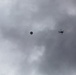 The image shows an overcast sky with two parachutes descending and a helicopter flying nearby. The silhouettes of the parachutes and helicopter are visible against a gray, cloudy background. This appears to depict a military or training airborne operation.