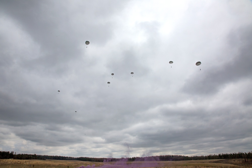 The image shows a military airborne operation with multiple paratroopers descending over an open field under overcast skies. Approximately seven to eight parachutes are visible at varying altitudes, suggesting a mass tactical jump. A tree line borders the landing zone, and a faint purple smoke signal is visible near the ground, likely used for wind direction or marking.