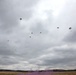 The image shows a military airborne operation with multiple paratroopers descending over an open field under overcast skies. Approximately seven to eight parachutes are visible at varying altitudes, suggesting a mass tactical jump. A tree line borders the landing zone, and a faint purple smoke signal is visible near the ground, likely used for wind direction or marking.