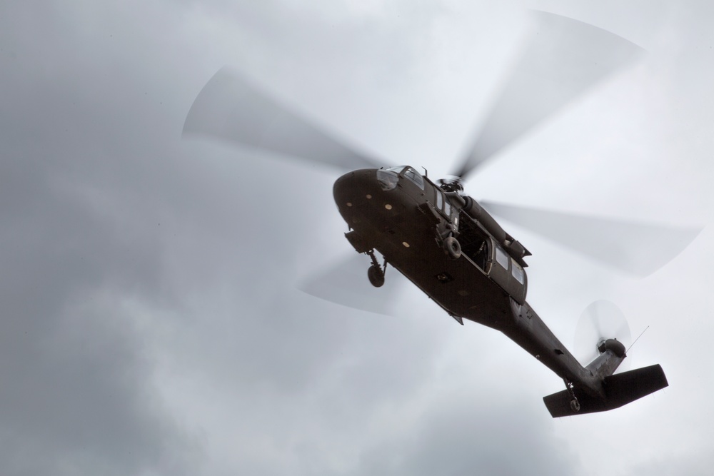A military helicopter is photographed from below against an overcast sky. The aircraft appears to be a utility/transport helicopter, likely a Sikorsky UH-60 Black Hawk, captured in flight with rotor blades in motion creating a motion blur effect. The image is taken from a low angle looking upward, providing a view of the helicopter's underside.